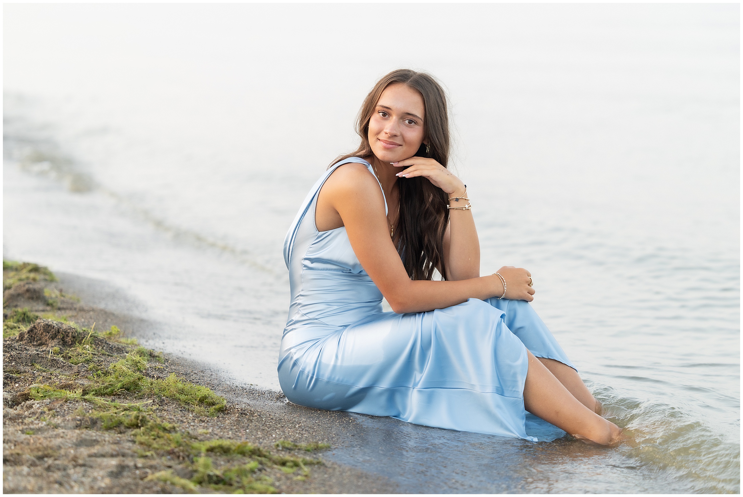 Young brunette woman sitting on the beach in a light blue satin dress for senior portraits