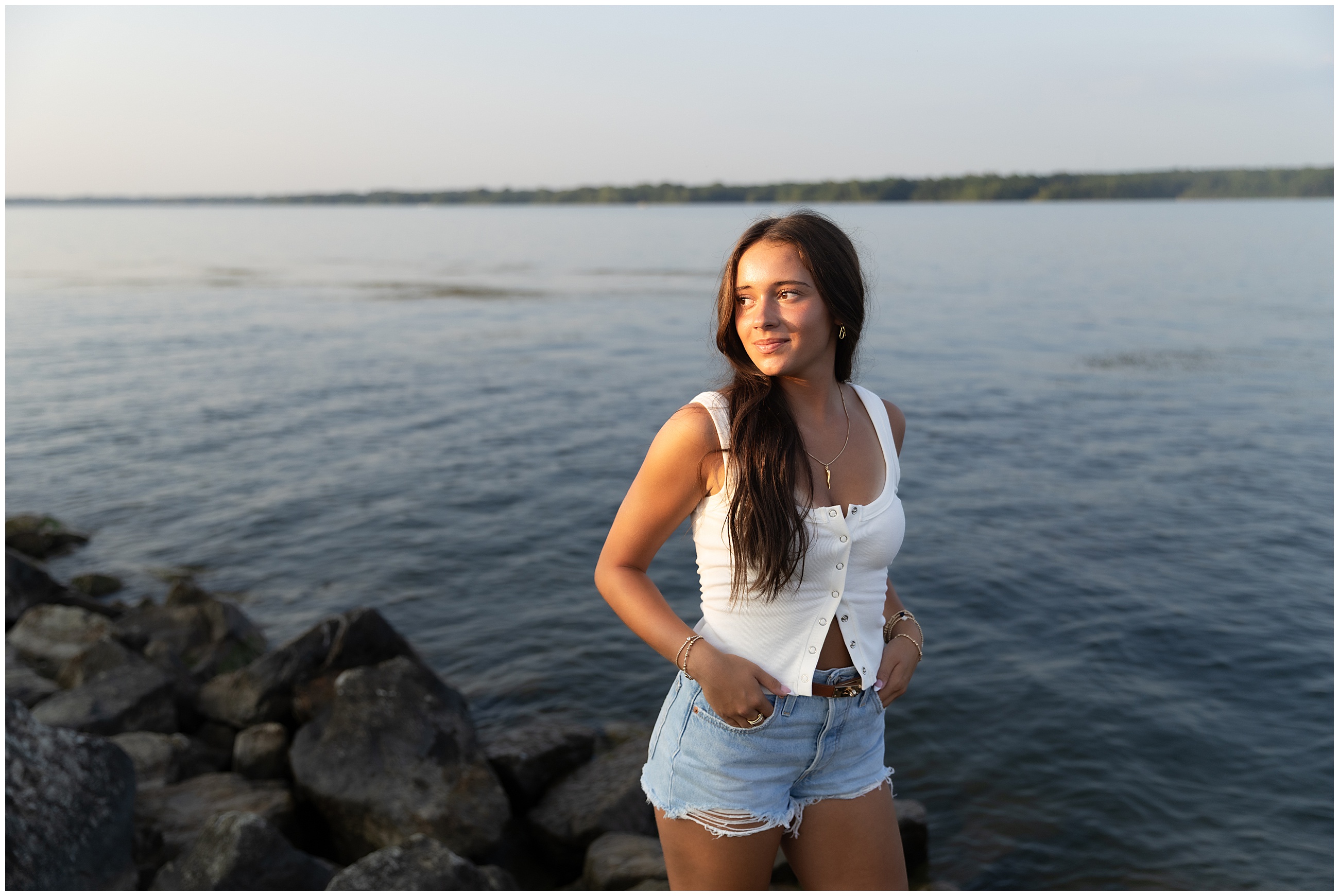 Young woman in a white top and denim shorts stands on rocky shore, smiling at sunset by a calm lake. Mood is serene and relaxed. High school senior portrait session.
