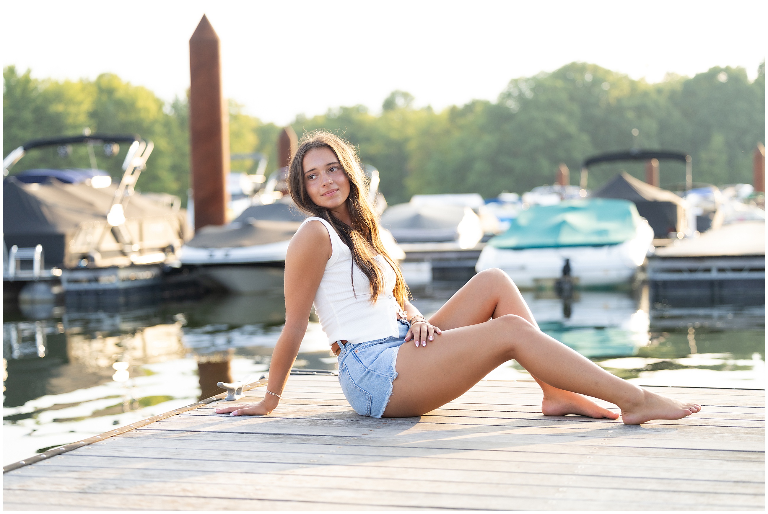 Woman in white top and denim shorts sits on a dock by the water with moored boats behind. Sunlit, serene atmosphere. High school senior portrait session.