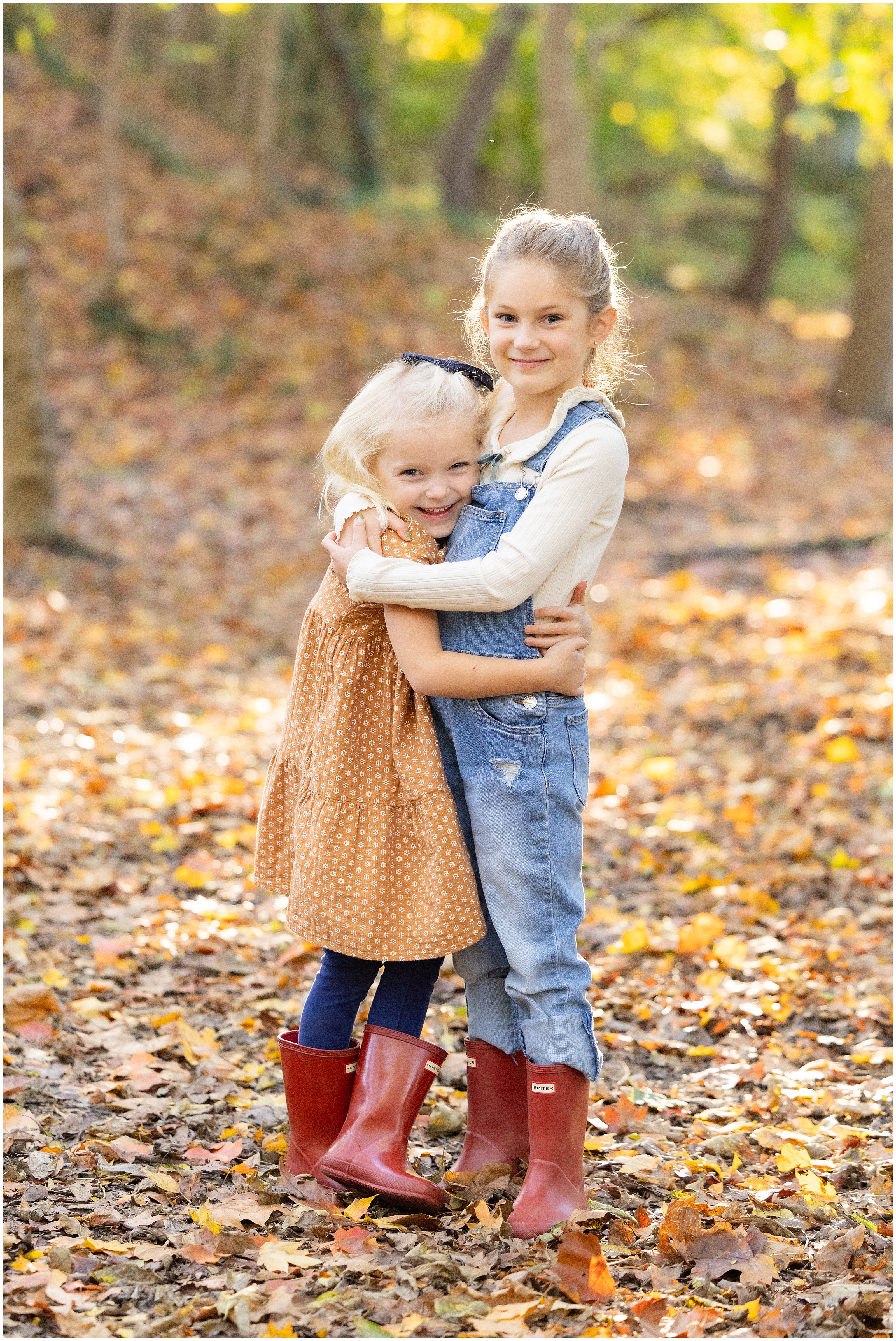 Two young girls hugging and smiling at the camera, wearing red rain boots, denim overalls and a dress, a colorful fall setting in the woods.