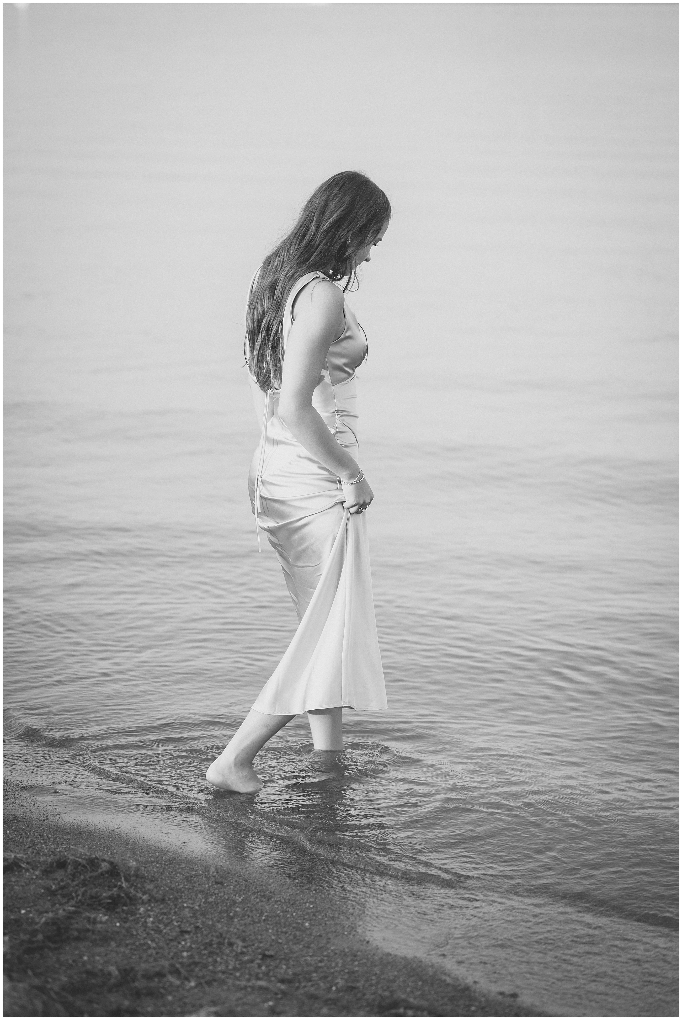 A black and white image of a young woman in a long dress walks barefoot along a calm shoreline. The scene is serene, with soft ripples in the water and a peaceful mood.